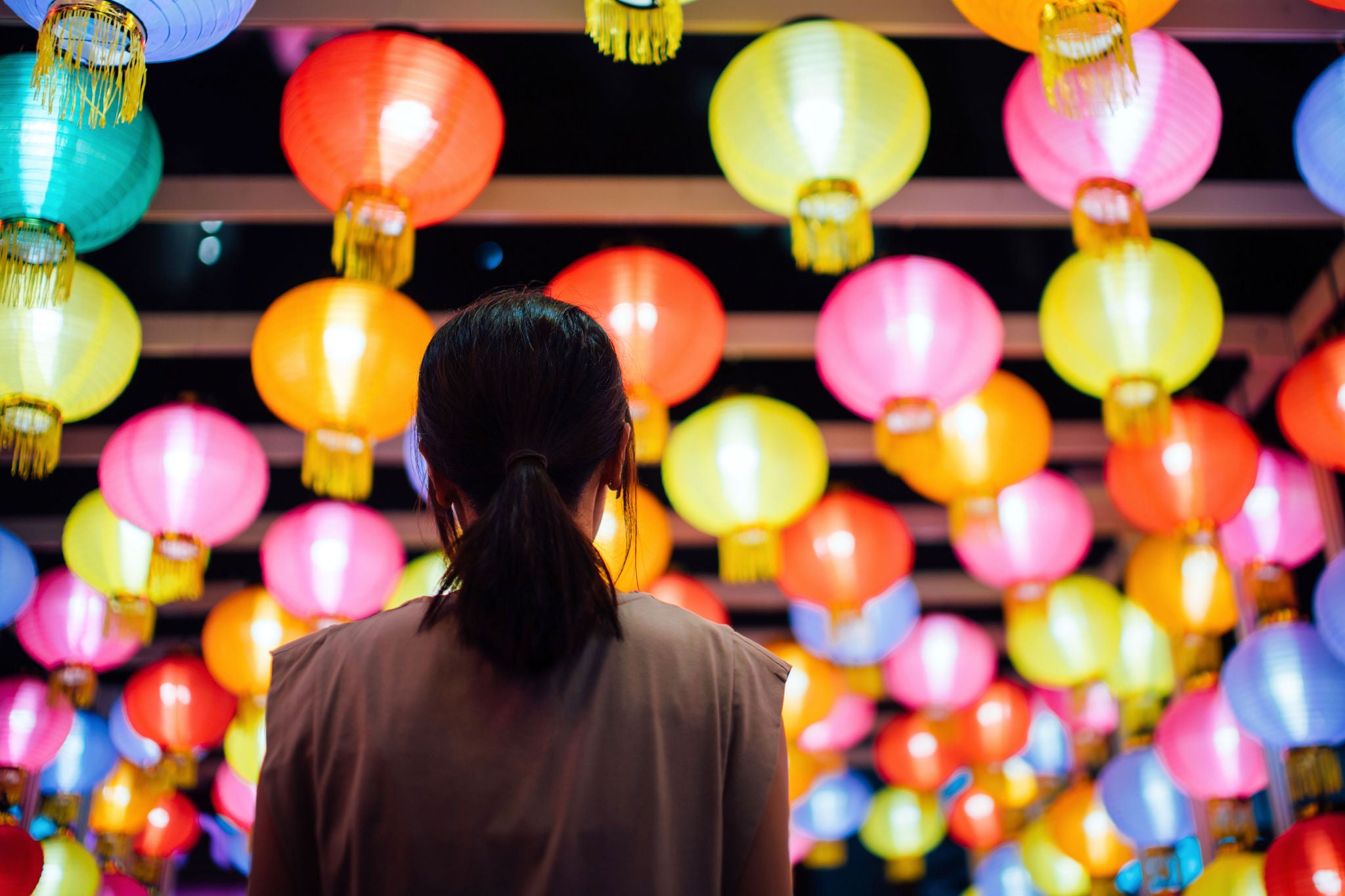 Rear view of young Asian woman looking up and admiring the hanging illuminated and colourful traditional Chinese lanterns on city street at night. Traditional Chinese culture, festival and celebration event theme